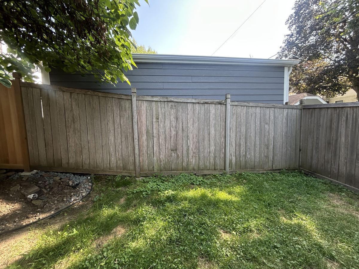 A privacy fence gone weathered grey-brown around a corner of a back yard, before restoration